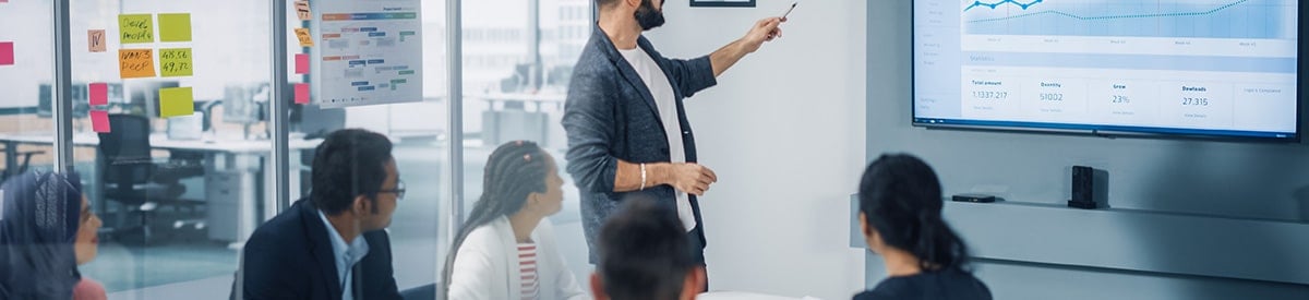 Isang lalaking naka-blazer ang nakatayo sa harap ng isang grupo ng mga kasamahan niya, nakaturo siya sa graph na naka-display sa malaking monitor